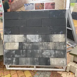 Ekspozycja kostki betonowej w odcieniach szarości, ułożonej w rzędach na stojaku, z widocznymi przebarwieniami i plamami na niektórych elementach, na tle wystawy innych materiałów budowlanych.