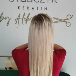 Tył głowy kobiety z długimi, prostymi, blond włosami po zabiegu keratynowym. Na ścianie logo salonu 'Stańczyk Keratin by Aleks' oraz złote nożyczki.