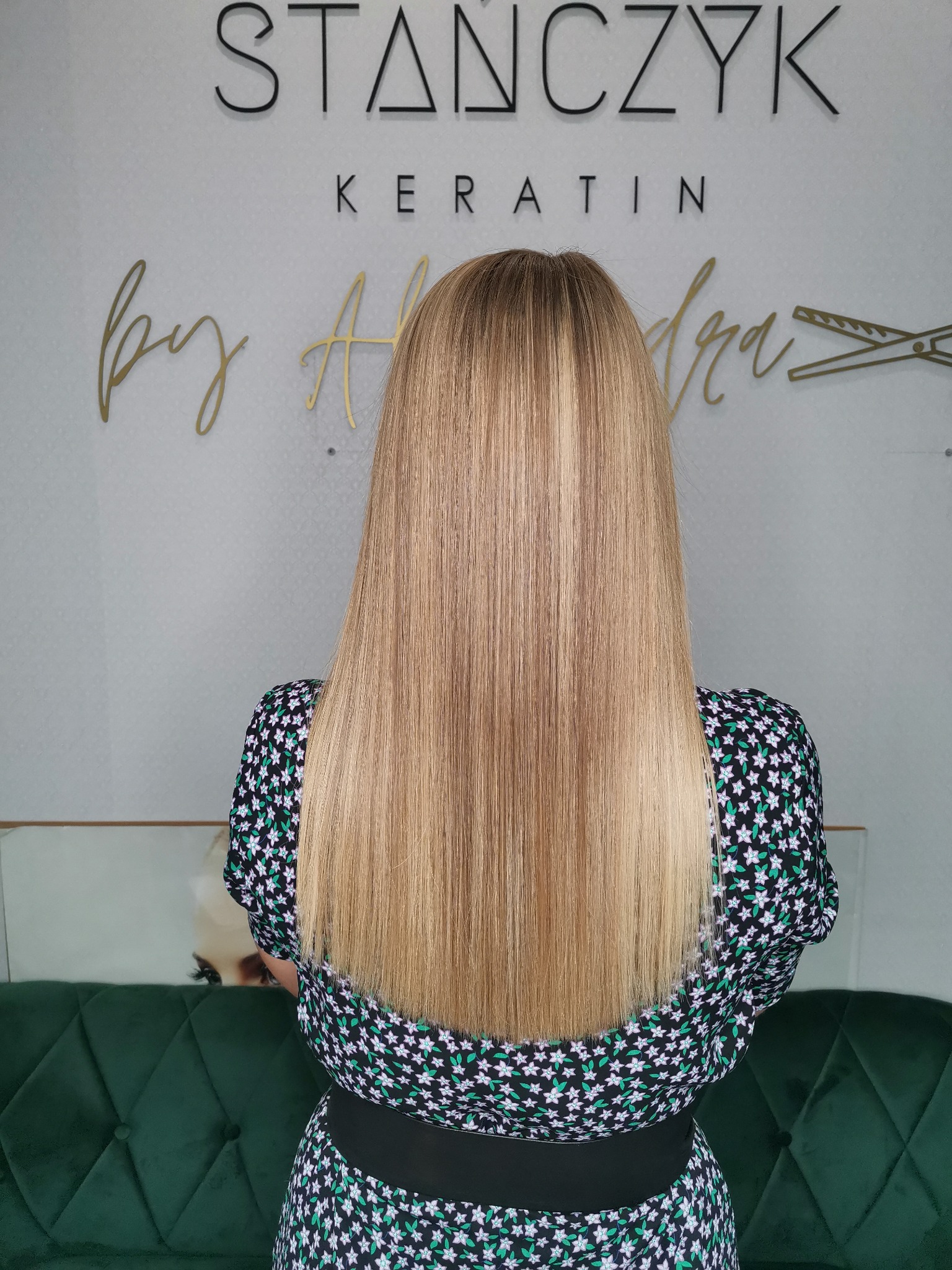 Proste, blond włosy z ciemniejszym pasemkiem, widok z tyłu, na tle ściany z napisem 'STAŃCZYK KERATIN by Aleksandra' i zielonej, welurowej kanapy.