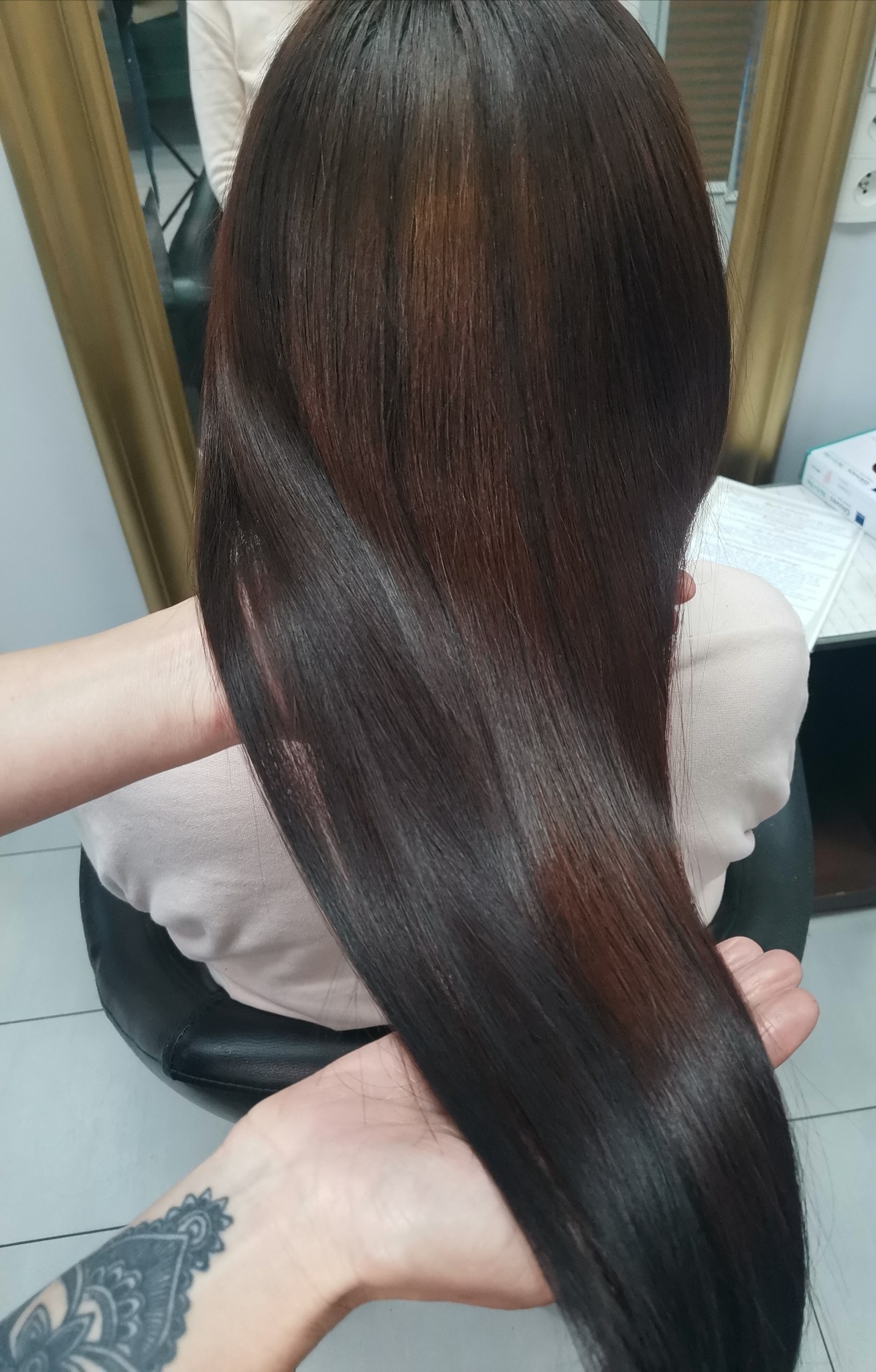 Długie, lśniące, brązowe włosy z refleksami, trzymane w dłoniach w salonie fryzjerskim, widoczny tatuaż na nadgarstku.