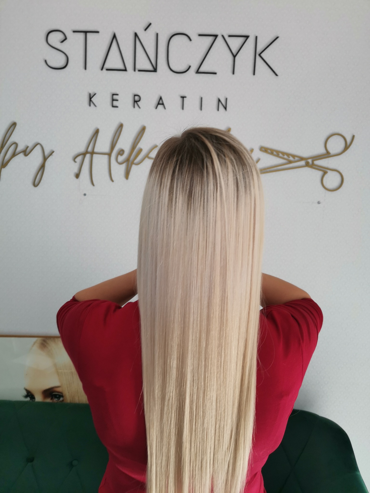 Tył głowy kobiety z długimi, prostymi, blond włosami po zabiegu keratynowym. Na ścianie logo salonu 'Stańczyk Keratin by Aleks' oraz złote nożyczki.