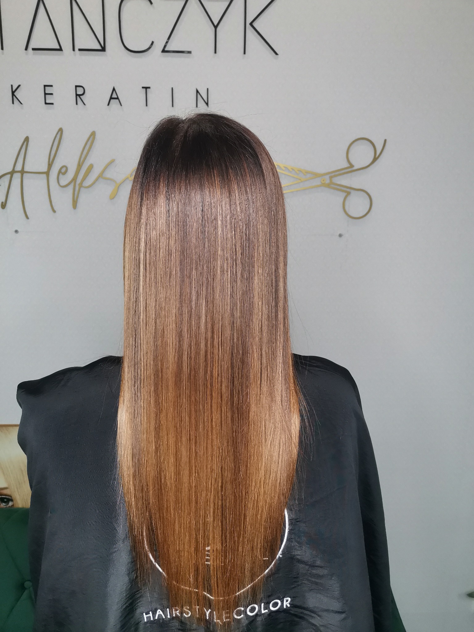 Proste, lśniące włosy po zabiegu keratynowym, z widocznym przejściem kolorów od ciemnego brązu u nasady do miedzianego blondu na końcach, na tle ściany z logo salonu.