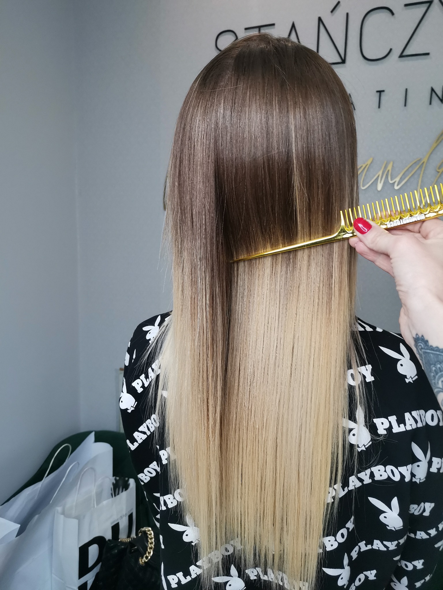 Rozjaśnione włosy klientki czesane złotym grzebieniem, widok z tyłu, na tle szaro-białego wnętrza salonu.