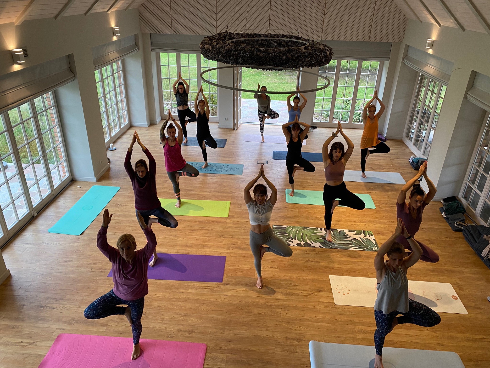 Grupowa sesja jogi w przestronnym, jasnym wnętrzu z dużymi oknami; uczestnicy w pozycji drzewa na kolorowych matach.