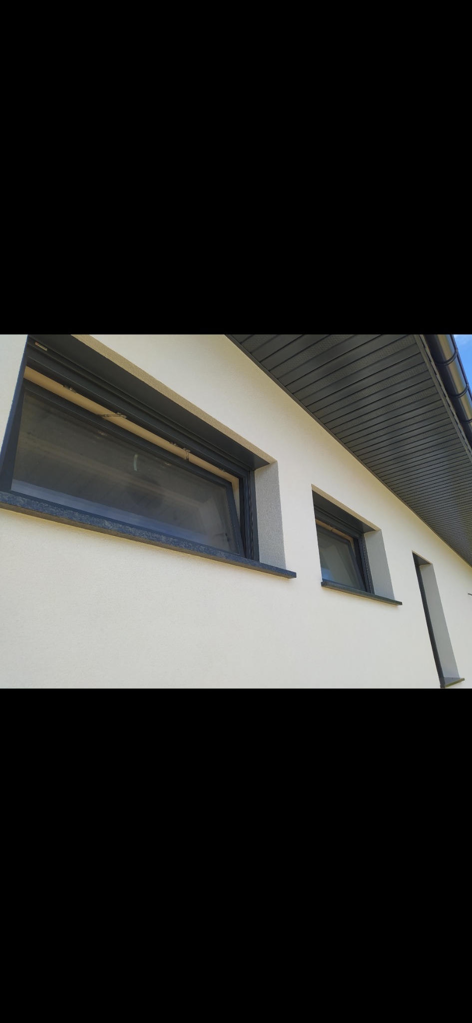 Jasna elewacja budynku z trzema oknami z czarnymi ramami i czarnymi parapetami, widoczny fragment podbitki dachowej