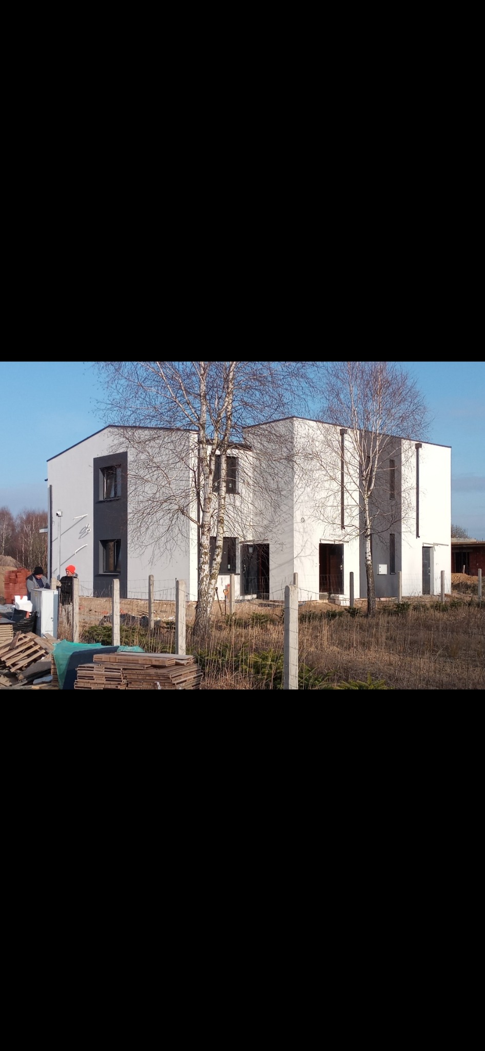 Dwukondygnacyjny budynek z elewacją w odcieniach szarości i bieli, widoczny zza betonowego ogrodzenia i dwóch brzóz, na tle niebieskiego nieba.