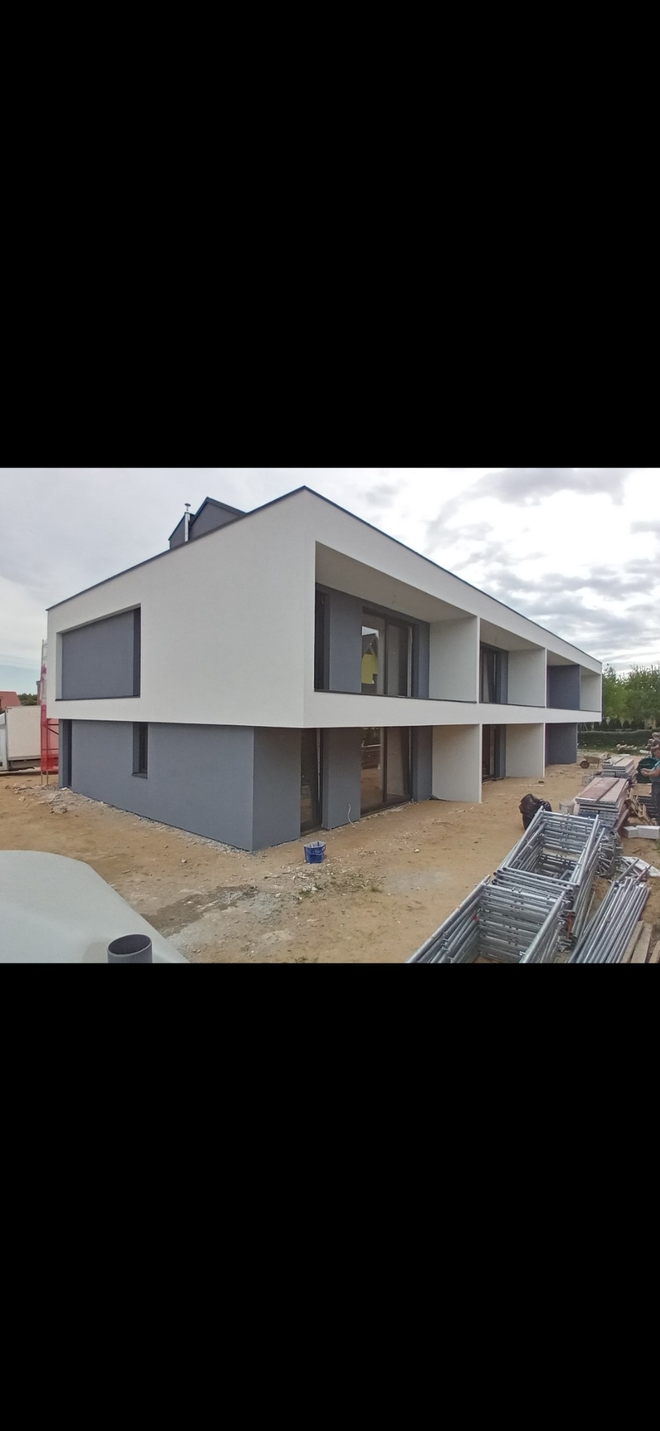 Nowoczesny, dwukondygnacyjny dom w trakcie budowy, z szarą dolną i białą górną częścią elewacji, widoczne rusztowania i teren budowy.