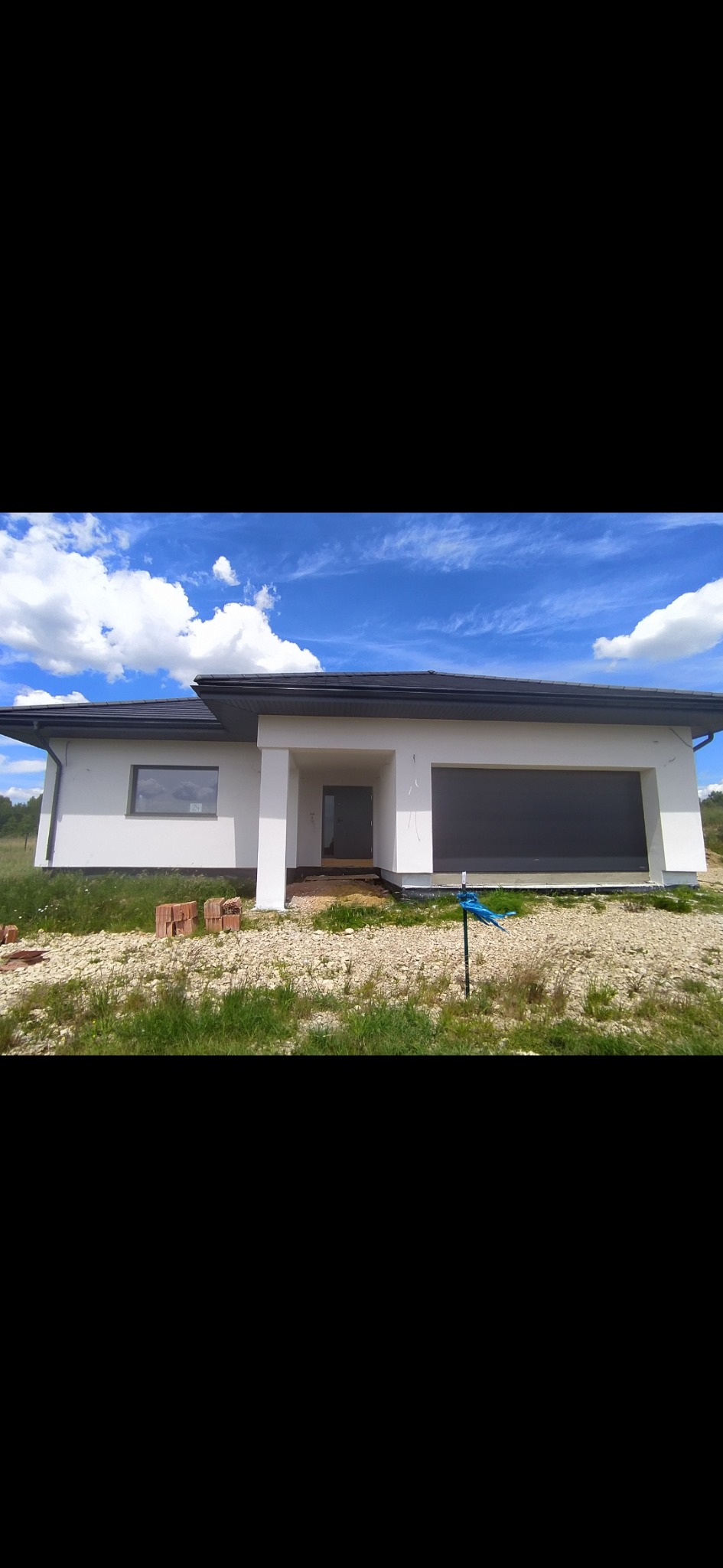 Nowoczesny dom jednorodzinny z białą elewacją i szarą bramą garażową, widok z zewnątrz na niezagospodarowaną działkę z gruzem i cegłami.