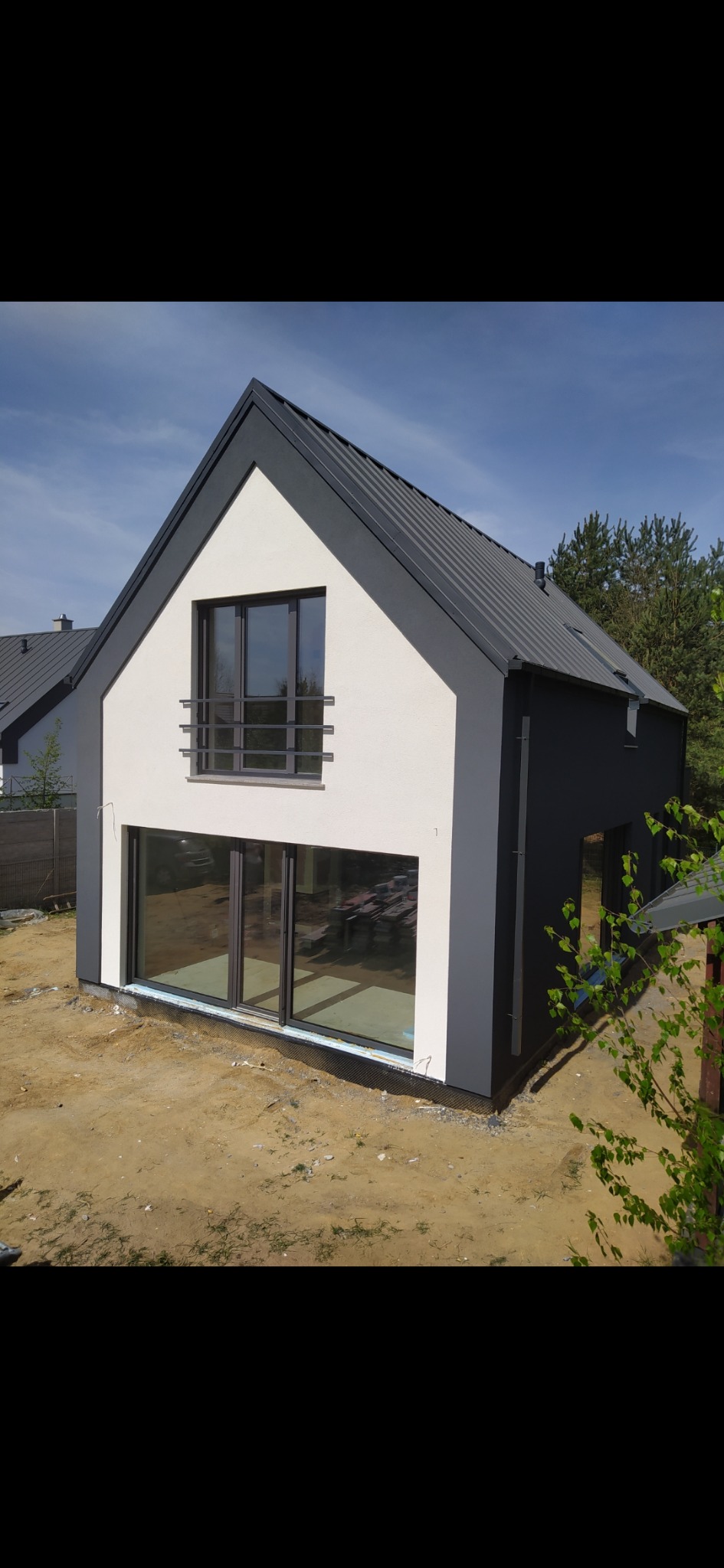 Nowoczesny dom jednorodzinny z elewacją w odcieniach szarości i bieli, z dużymi oknami i metalową balustradą, widoczny na tle nieba i drzew, teren wokół domu w trakcie zagospodarowywania.