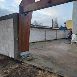 Mar mat-bud-instal - Wznoszenie ściany z bloczków betonowych; widoczny fragment konstrukcji drewnianej bramy wjazdowej, z tyłu komin fabryczny.