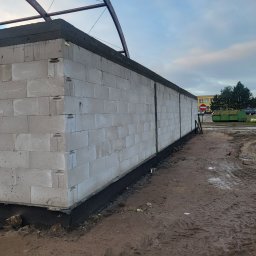 Mar mat-bud-instal - Ściana domu w trakcie budowy z betonu komórkowego, widoczne izolacja fundamentów i zadaszenie w surowym stanie, teren budowy błotnisty.