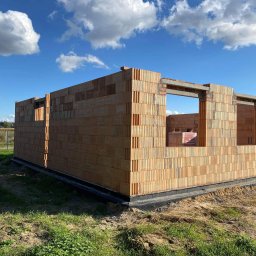 Mar mat-bud-instal - Wznoszone ściany domu z pustaków ceramicznych Porotherm, widoczna hydroizolacja fundamentów i fragment drugiego budynku w tle, błękitne niebo z chmurami.