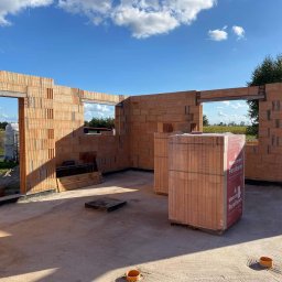 Mar mat-bud-instal - Wznoszenie ścian parteru domu z bloczków ceramicznych Porotherm, widoczne otwory okienne i drzwiowe, palety z materiałem budowlanym wewnątrz budynku, błękitne niebo w tle.