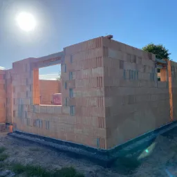 Wznoszone ściany parterowego domu z pustaków ceramicznych Porotherm, widoczna izolacja fundamentów i paleta pustaków obok otworu okiennego, słoneczny dzień.