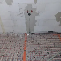 Montaż ogrzewania podłogowego w nowym budynku z bloków betonu komórkowego; widoczne rury grzewcze mocowane do maty z folią aluminiową, ściana w stanie surowym z widocznymi śladami po instalacjach.