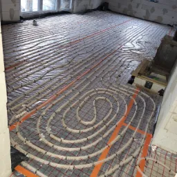 Układanie ogrzewania podłogowego w surowym wnętrzu, rury grzewcze ułożone na folii z siatką, widoczne okna i ściany z bloczków.