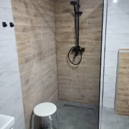 Wykończona kabina prysznicowa z czarną armaturą, wyłożona płytkami imitującymi drewno, z małym stołkiem ułatwiającym korzystanie.