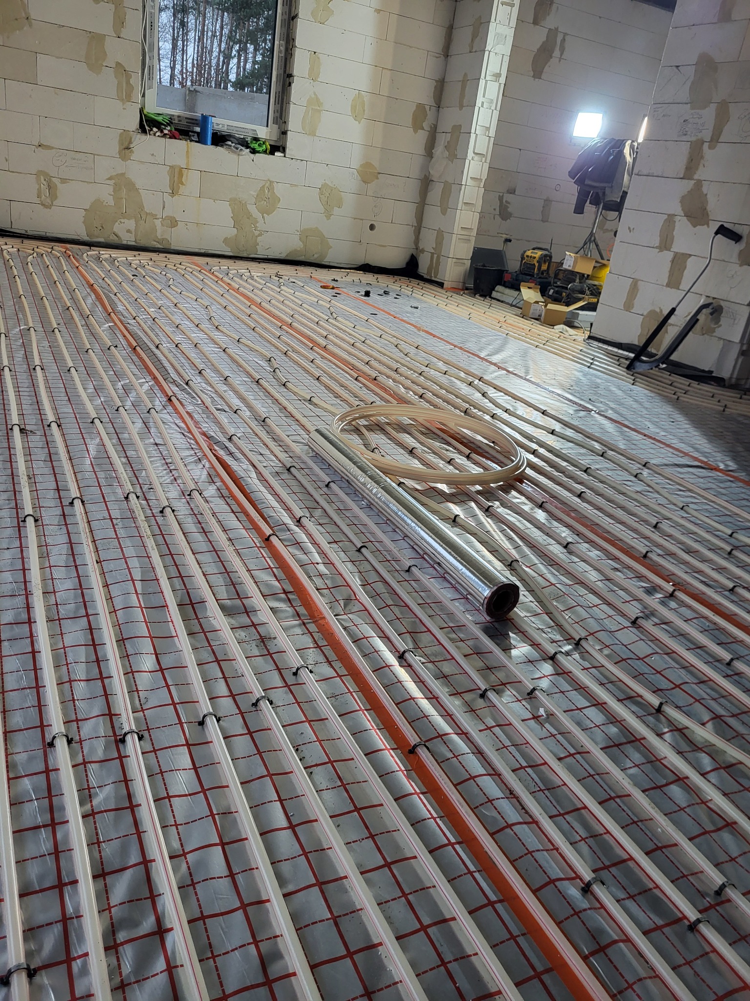 Układanie ogrzewania podłogowego w surowym stanie budynku: rury grzewcze rozłożone na folii z siatką, widoczne narzędzia i sprzęt budowlany w tle.