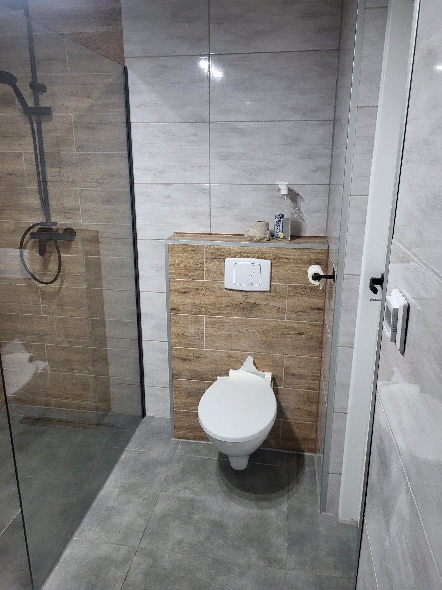 Nowoczesna łazienka z kabiną prysznicową typu walk-in, podwieszaną toaletą z zabudową w imitacji drewna i szarą płytką podłogową.