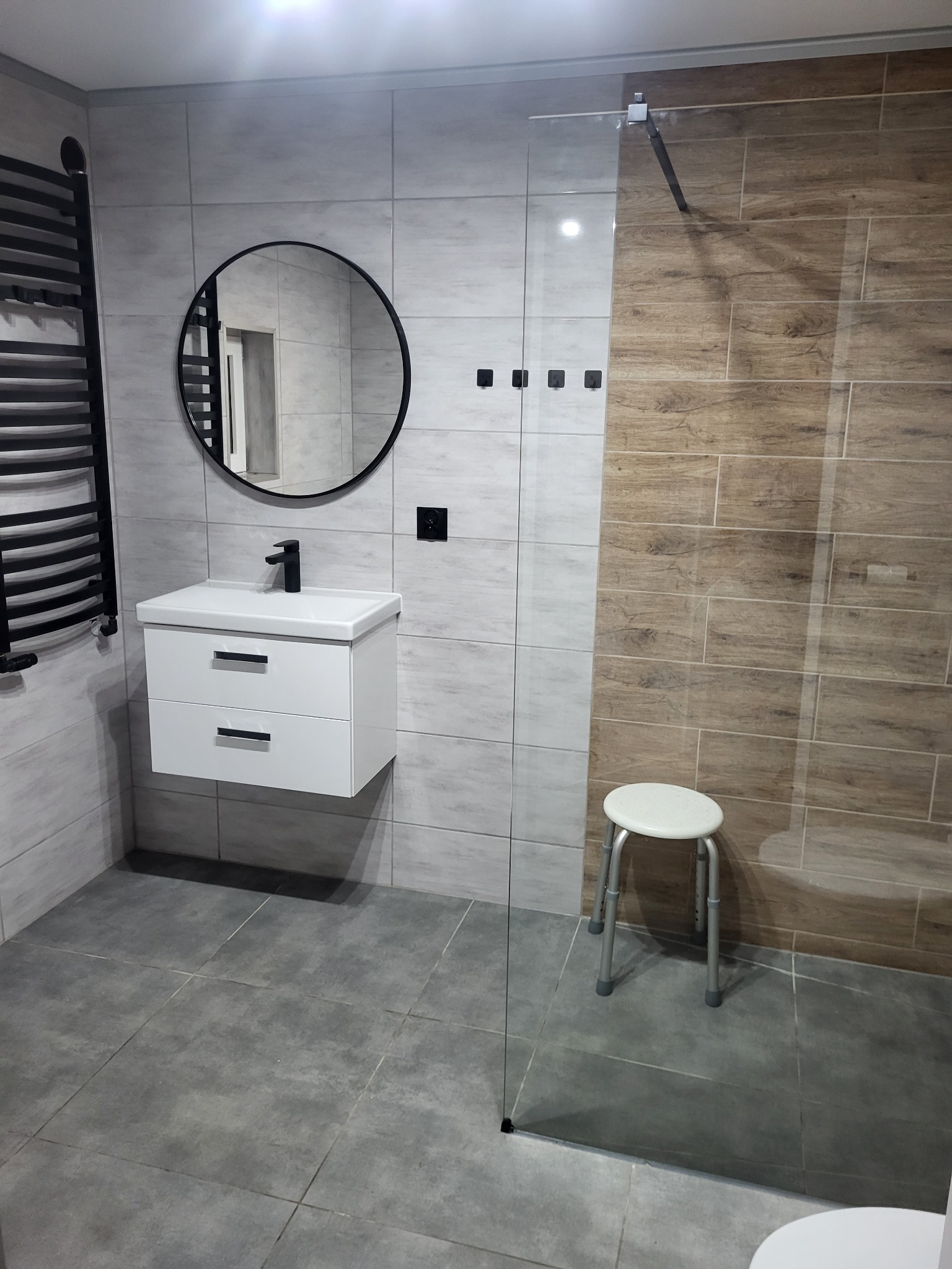 Nowoczesna łazienka z szarymi i drewnopodobnymi płytkami, okrągłym lustrem nad białą szafką z umywalką i szklaną kabiną prysznicową.