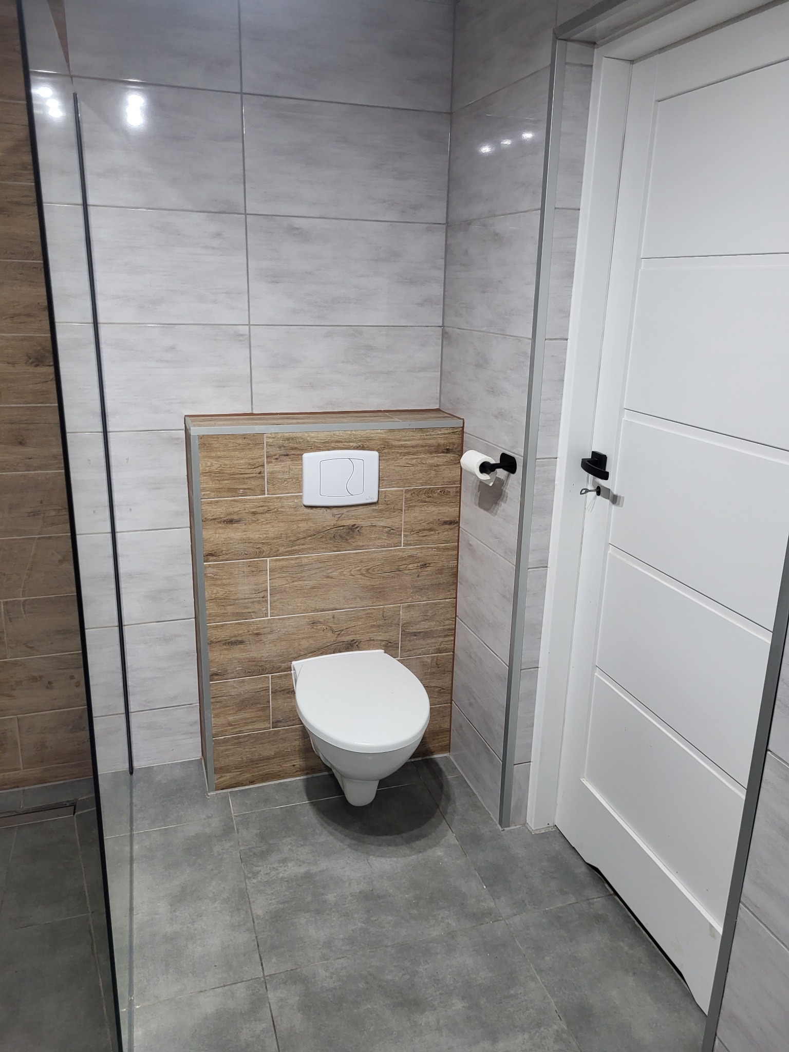 Nowoczesna toaleta podwieszana z zabudową z płytek imitujących drewno, szare płytki podłogowe, białe płytki ścienne, białe drzwi z czarną klamką i uchwyt na papier toaletowy.