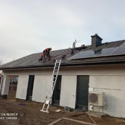 Instalator z zabezpieczeniami na dachu domu jednorodzinnego montuje panele słoneczne; widoczna drabina, antena satelitarna i jednostka zewnętrzna pompy ciepła.