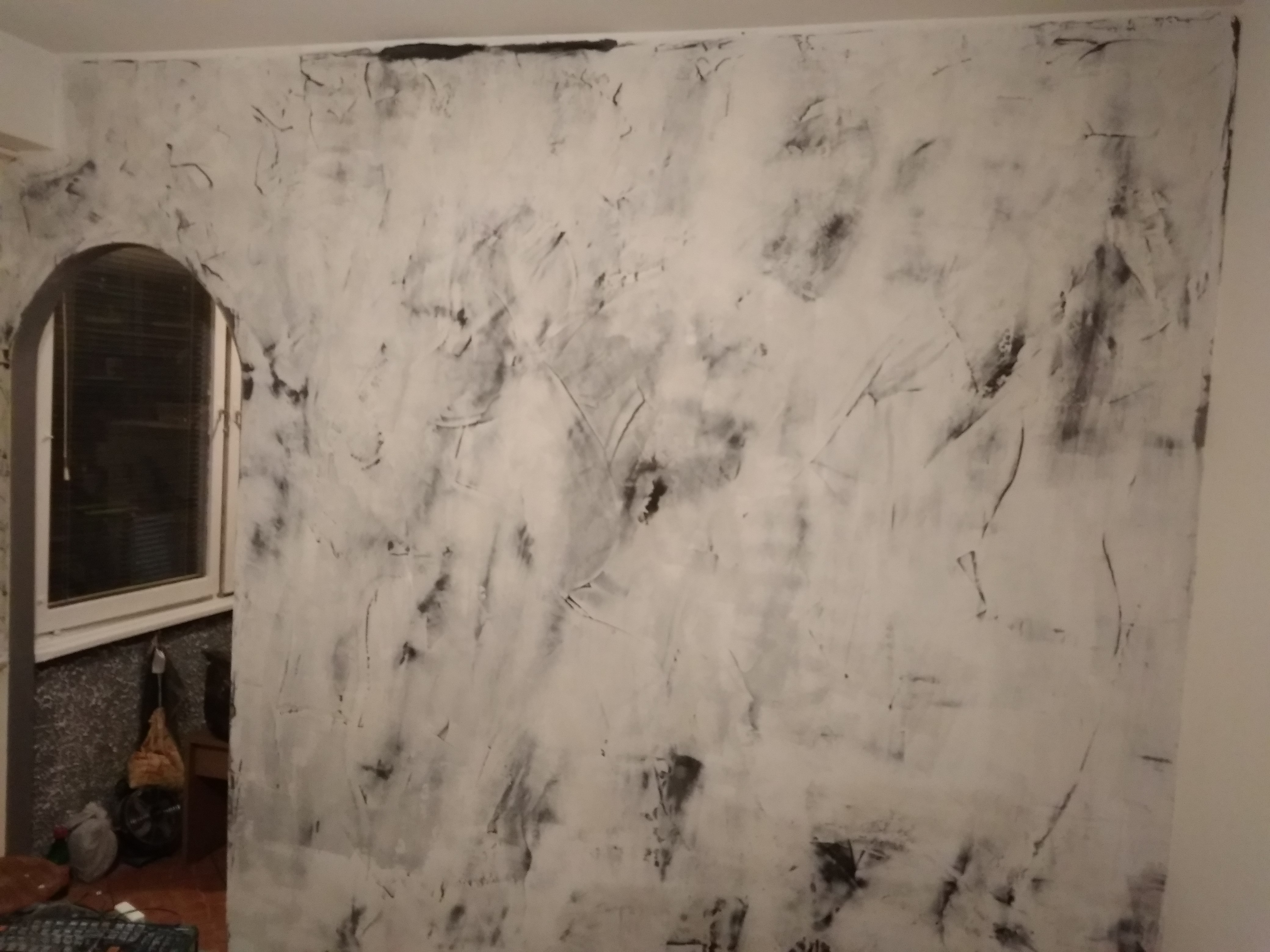 Ściana w trakcie szpachlowania z widocznymi smugami szpachli w odcieniach szarości i bieli, obok okno z widocznym parapetem i elementami wyposażenia wnętrza.