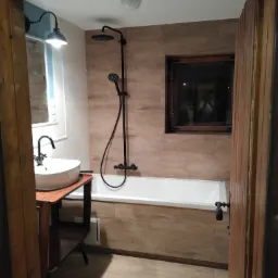 Wykończona łazienka z wanną, umywalką na drewnianym stelażu i czarną armaturą, widoczna przez otwarte drewniane drzwi.