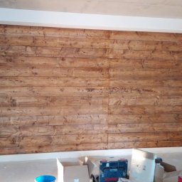 Edmar - Ściana wykończona panelami z surowego, impregnowanego drewna w odcieniach brązu, widoczne narzędzia i materiały budowlane w nieuporządkowanym otoczeniu podczas prac wykończeniowych.