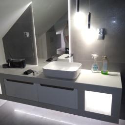 Edmar - Nowoczesna łazienka w szarościach z podświetlanym blatem pod umywalką i lustrem, widoczne butelki z płynami i radio na blacie, sufit ze skosem.