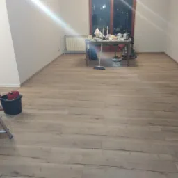 Ułożone panele podłogowe w pokoju z widocznym stołem roboczym, wiadrem i drabiną malarską w tle.