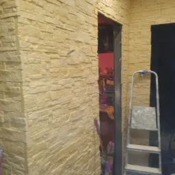 Wykończona ściana z dekoracyjnej, jasnej cegły, widoczna drabina malarska i wałek malarski z fioletową rączką, wnętrze w trakcie prac wykończeniowych.