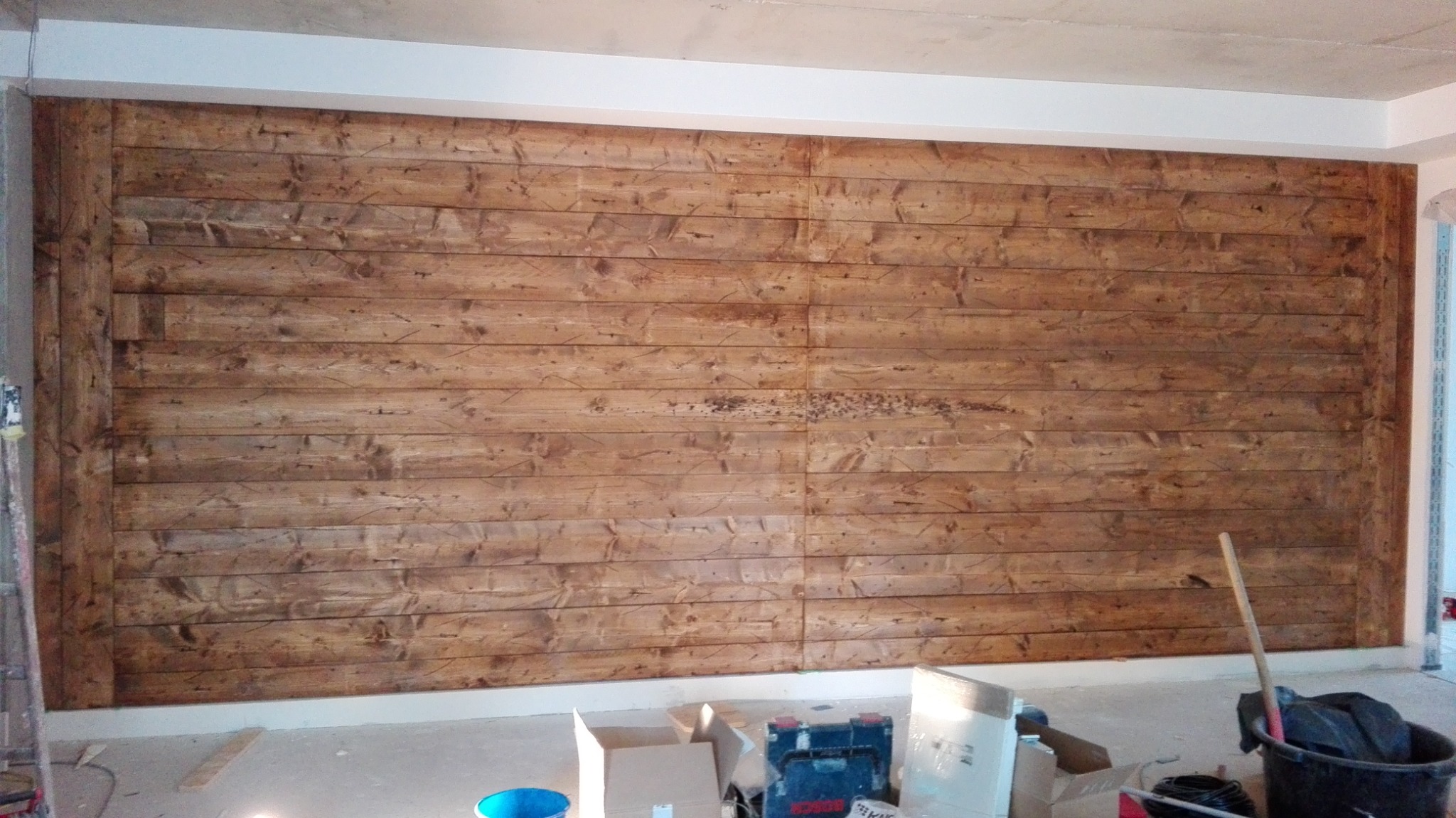 Ściana wykończona panelami z surowego, impregnowanego drewna w odcieniach brązu, widoczne narzędzia i materiały budowlane w nieuporządkowanym otoczeniu podczas prac wykończeniowych.