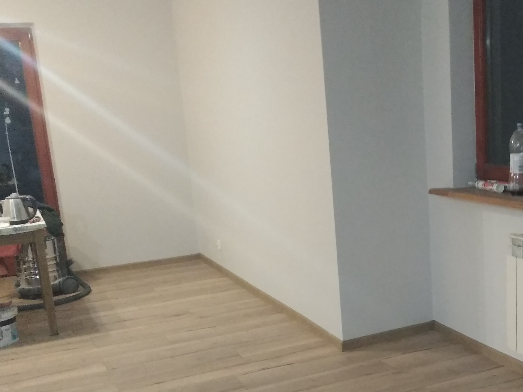 Wnętrze pokoju w trakcie prac wykończeniowych, widoczne świeżo pomalowane ściany w odcieniach bieli i szarości, drewniana podłoga, grzejnik oraz sprzęt do sprzątania.