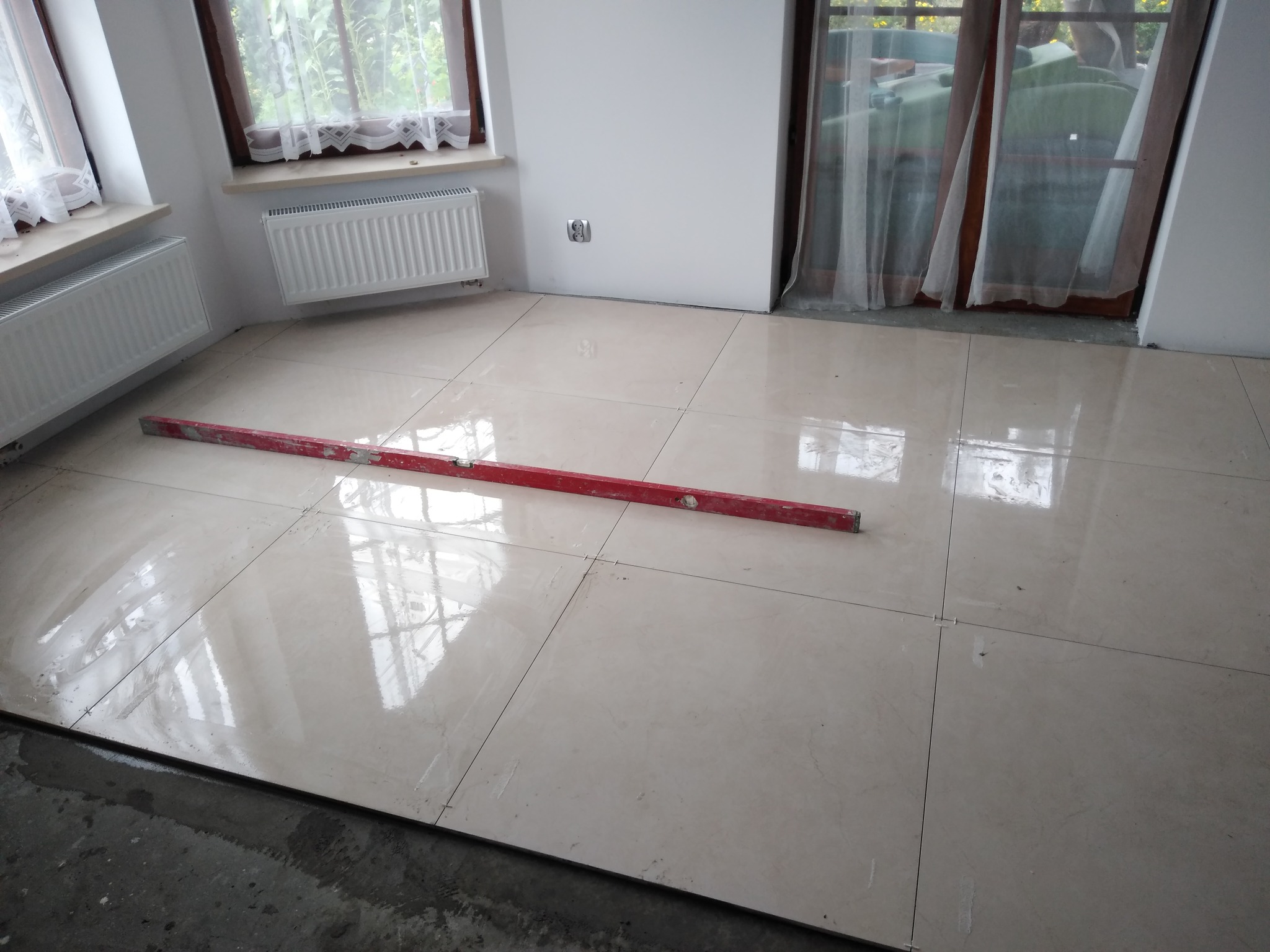 Jasne, błyszczące płytki podłogowe, częściowo ułożone, z widocznymi liniami fug i poziomicą w kolorze czerwonym, okna z białymi firankami i grzejniki w tle.