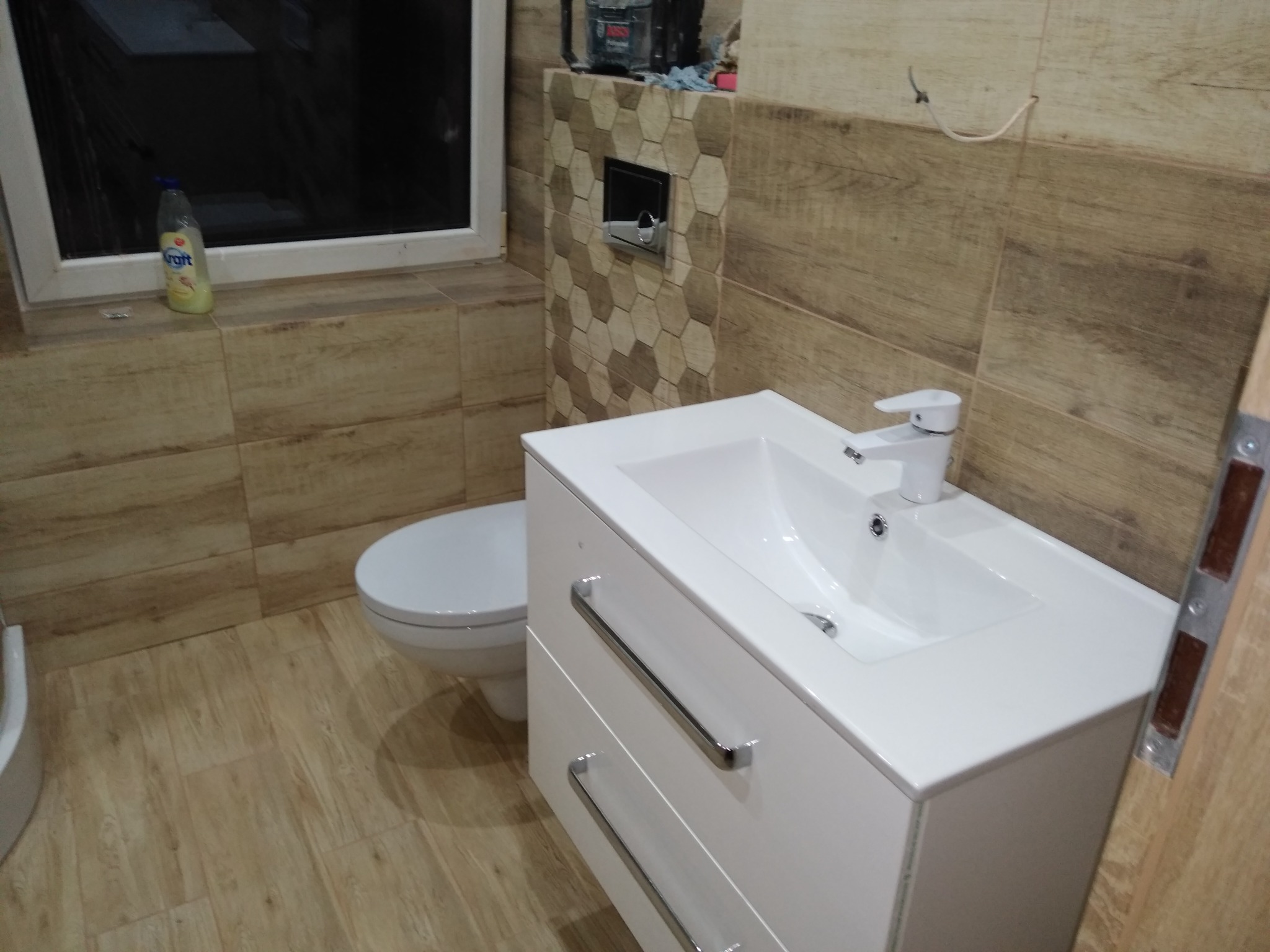 Wykończona łazienka z drewnopodobnymi płytkami, białą szafką z umywalką i wiszącą toaletą, widoczna butelka płynu do mycia okien na parapecie.