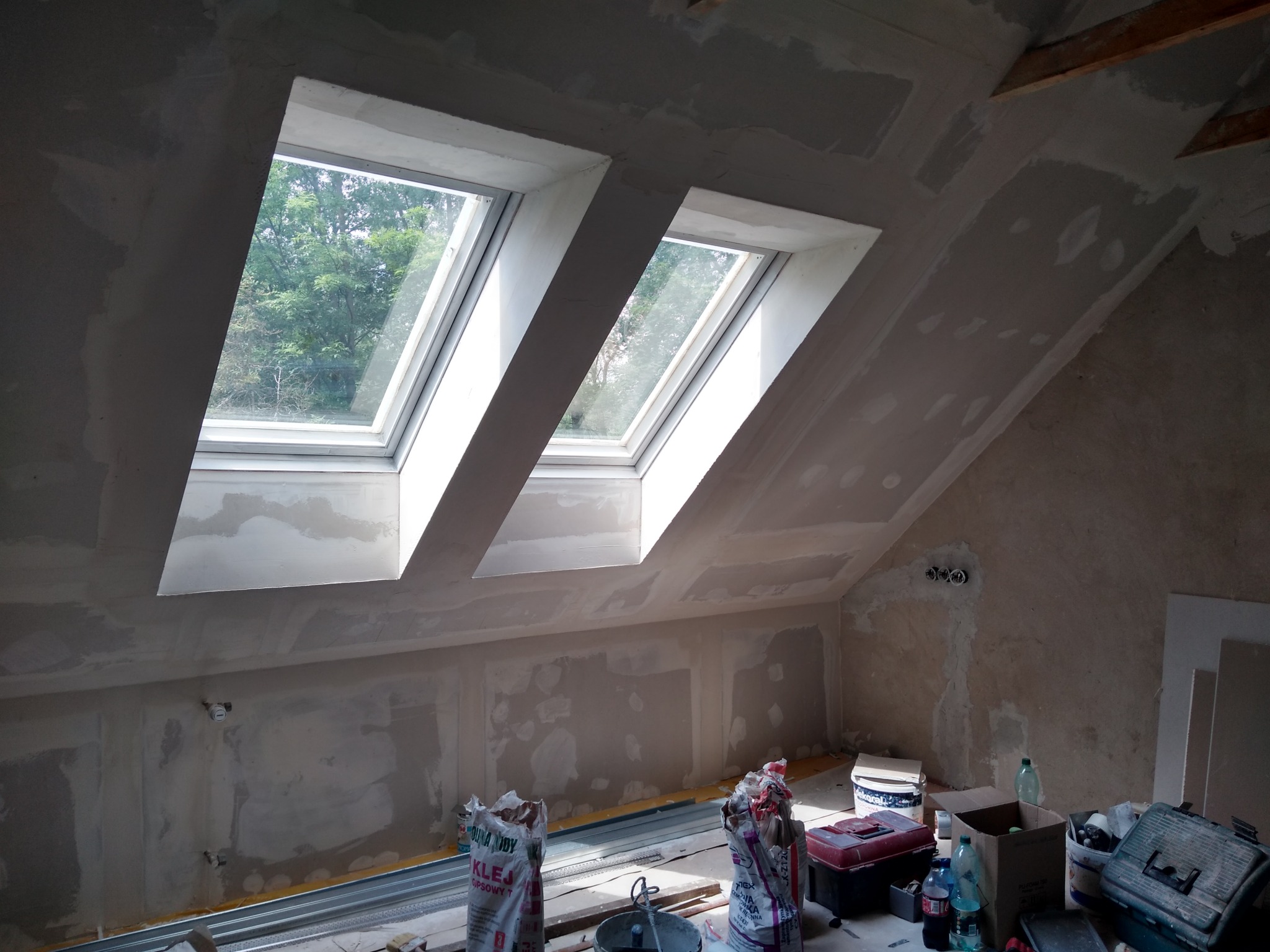 Poddasze w trakcie prac wykończeniowych, widoczne dwa okna dachowe, ściany przygotowane do malowania, worki z klejem gipsowym i narzędzia w nieładzie.