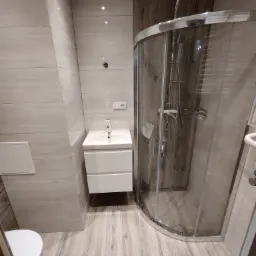 Nowoczesna łazienka z kabiną prysznicową z chromowanymi detalami, umywalką z szafką w kolorze białym i płytkami imitującymi drewno na podłodze i ścianach.