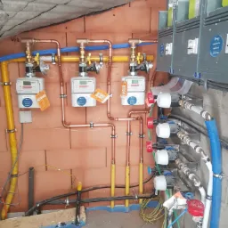 Instalacja gazowa i wodna w surowym pomieszczeniu z widocznymi licznikami, rurami miedzianymi i ż&oacute;łtymi, oraz elementami sterującymi, zamontowana na ceglanej ścianie i szarej ścianie z widocznymi...