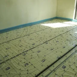 Białe rurki ogrzewania podłogowego ułożone na izolacji z niebieską folią przy ścianach, widoczne czarne klipsy mocujące i logo 'BEGETUBE' na macie izolacyjnej, przygotowane do wylewki.