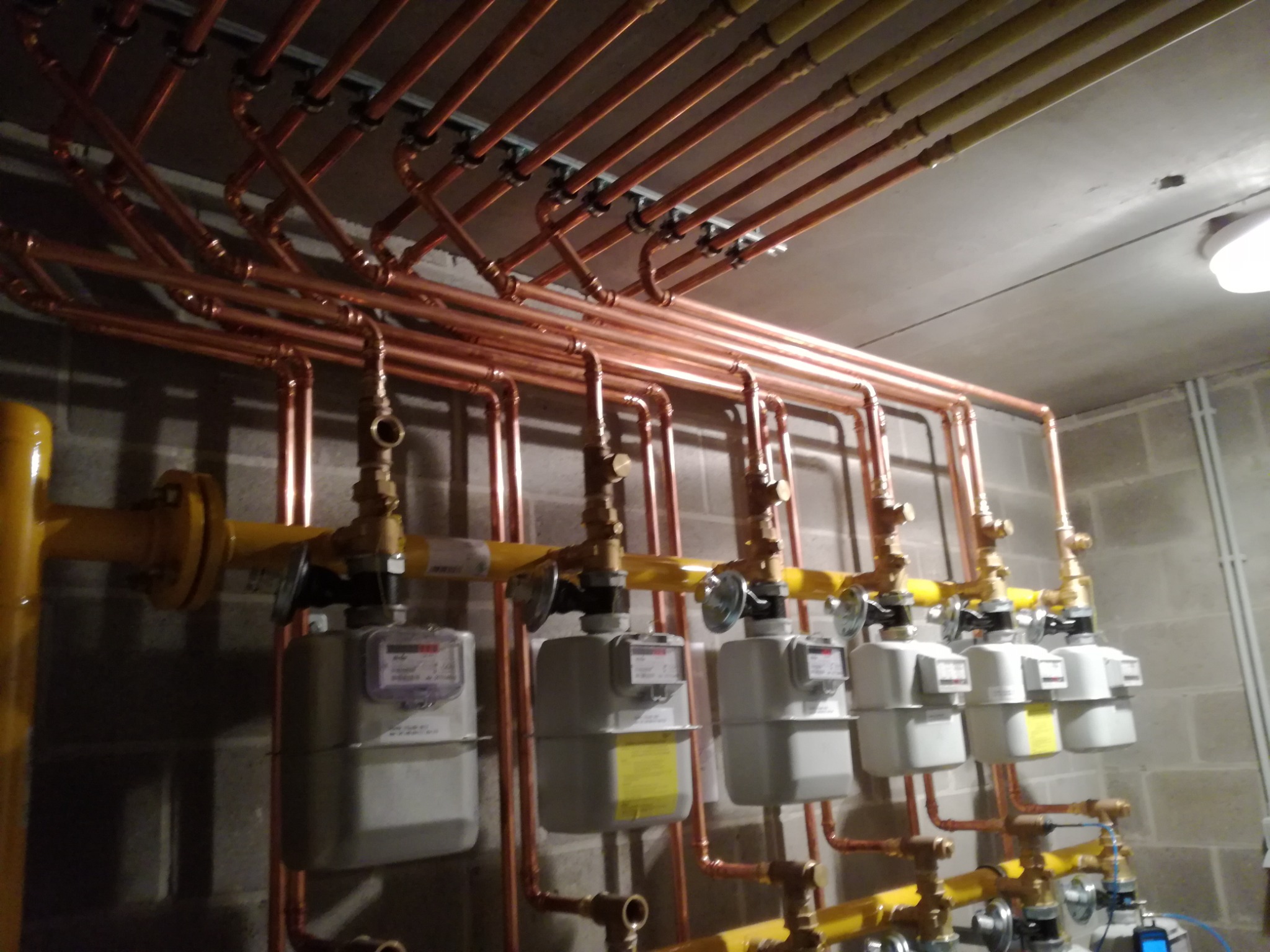 Skomplikowany system rur miedzianych i żółtych stalowych rur gazowych z zamontowanymi gazomierzami w pomieszczeniu o betonowej ścianie z bloków i suficie. Rury poprowadzone równolegle pod sufitem...