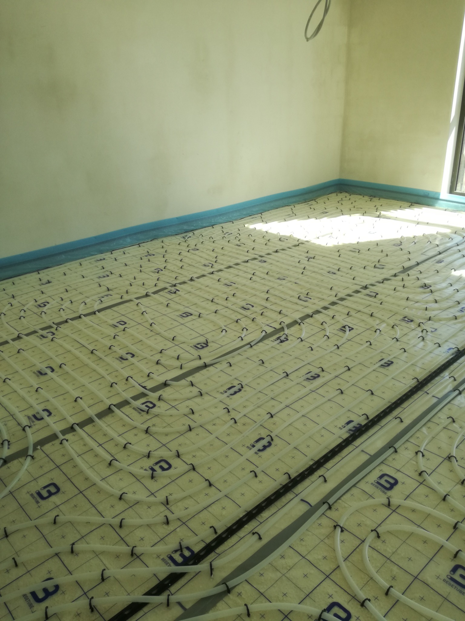 Białe rurki ogrzewania podłogowego ułożone na izolacji z niebieską folią przy ścianach, widoczne czarne klipsy mocujące i logo 'BEGETUBE' na macie izolacyjnej, przygotowane do wylewki.