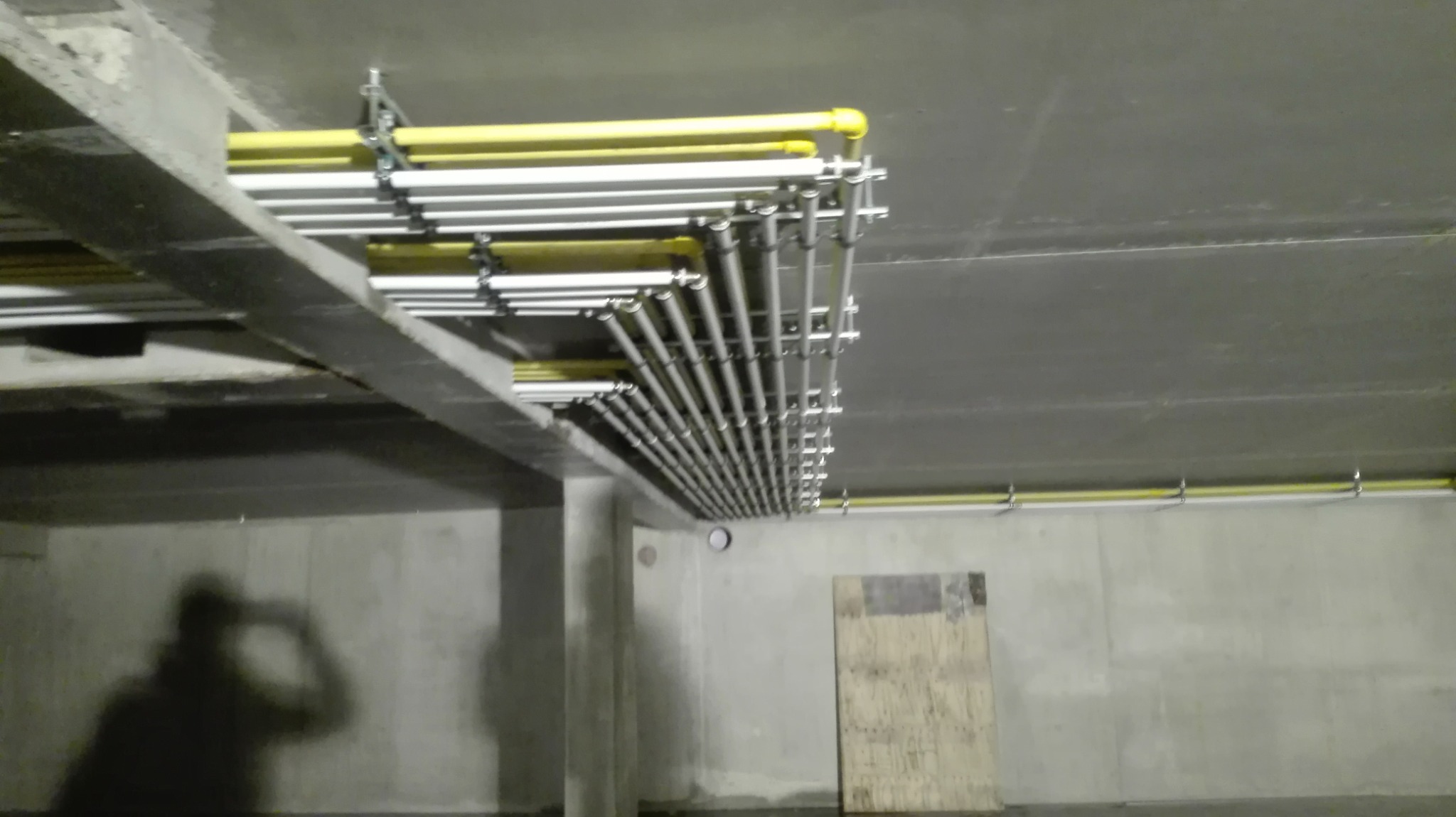 Równoległe rzędy rur instalacyjnych, częściowo żółtych, częściowo srebrnych, zamocowanych pod betonowym sufitem w surowym pomieszczeniu. Widoczny cień osoby na ścianie.
