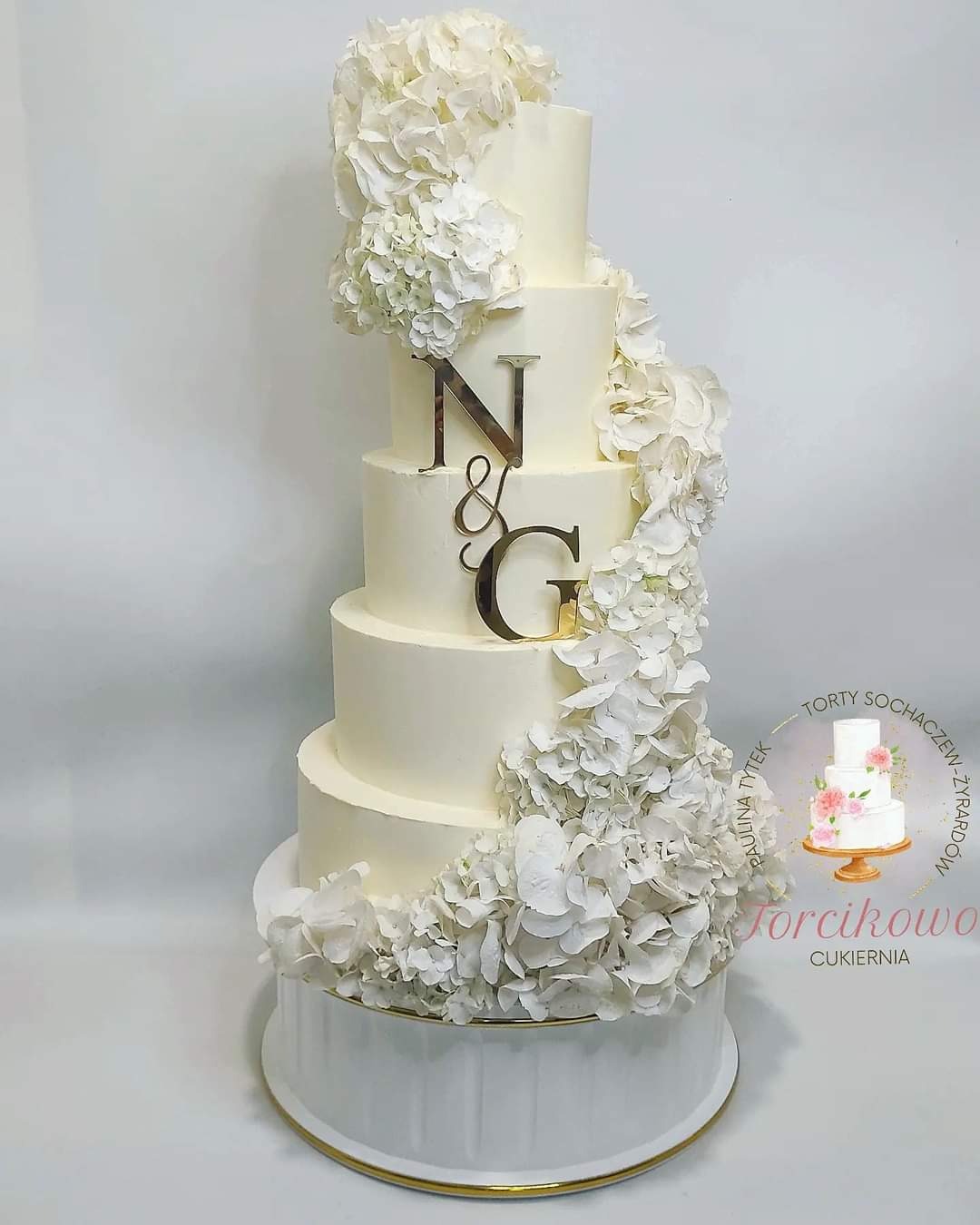 Elegancki, pięciopiętrowy tort weselny w odcieniach bieli, udekorowany kaskadą białych hortensji i złotymi inicjałami 'N & G', umieszczony na białej podstawie z delikatnym złotym akcentem.