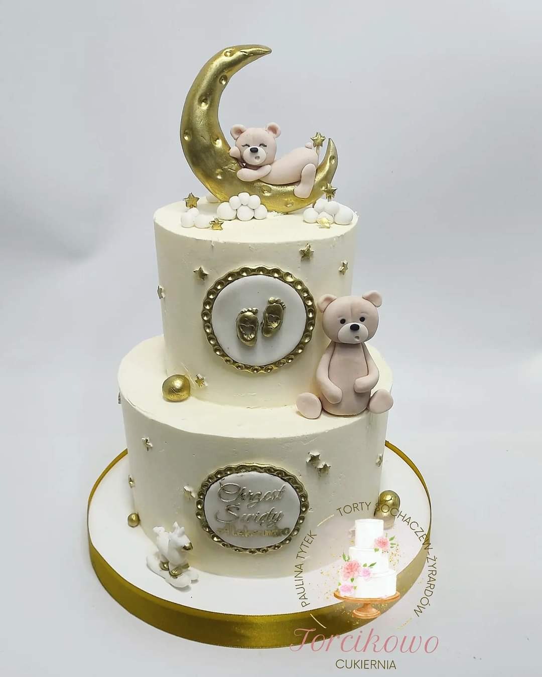 Dwupiętrowy tort na chrzest z dekoracjami: śpiący miś na złotym księżycu, złote stópki, małe gwiazdki i imię dziecka, umieszczony na złotym podkładzie.