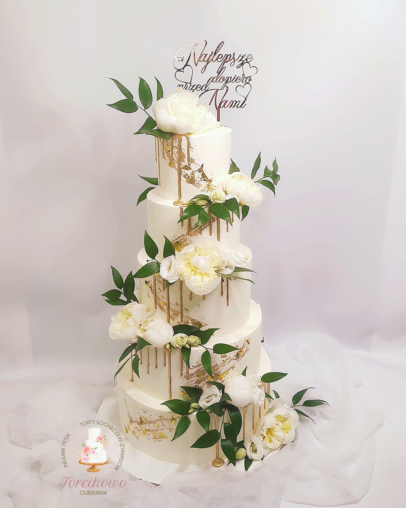 Elegancki, piętrowy tort weselny z białym kremem, złotymi akcentami i dekoracjami z żywych, białych kwiatów i zielonych liści, zwieńczony topperem z napisem.
