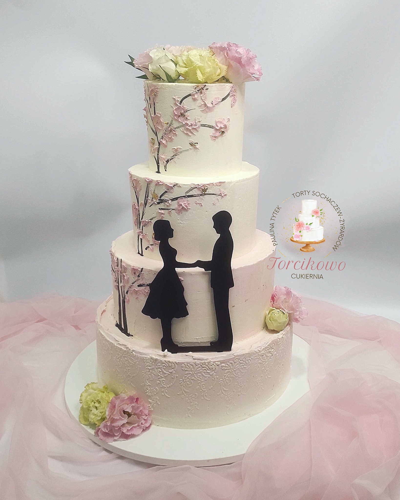Elegancki, czteropiętrowy tort weselny z motywem kwiatów wiśni i sylwetkami pary młodej, wykonany przez cukiernię z Sochaczewa, na delikatnym, różowym tle.