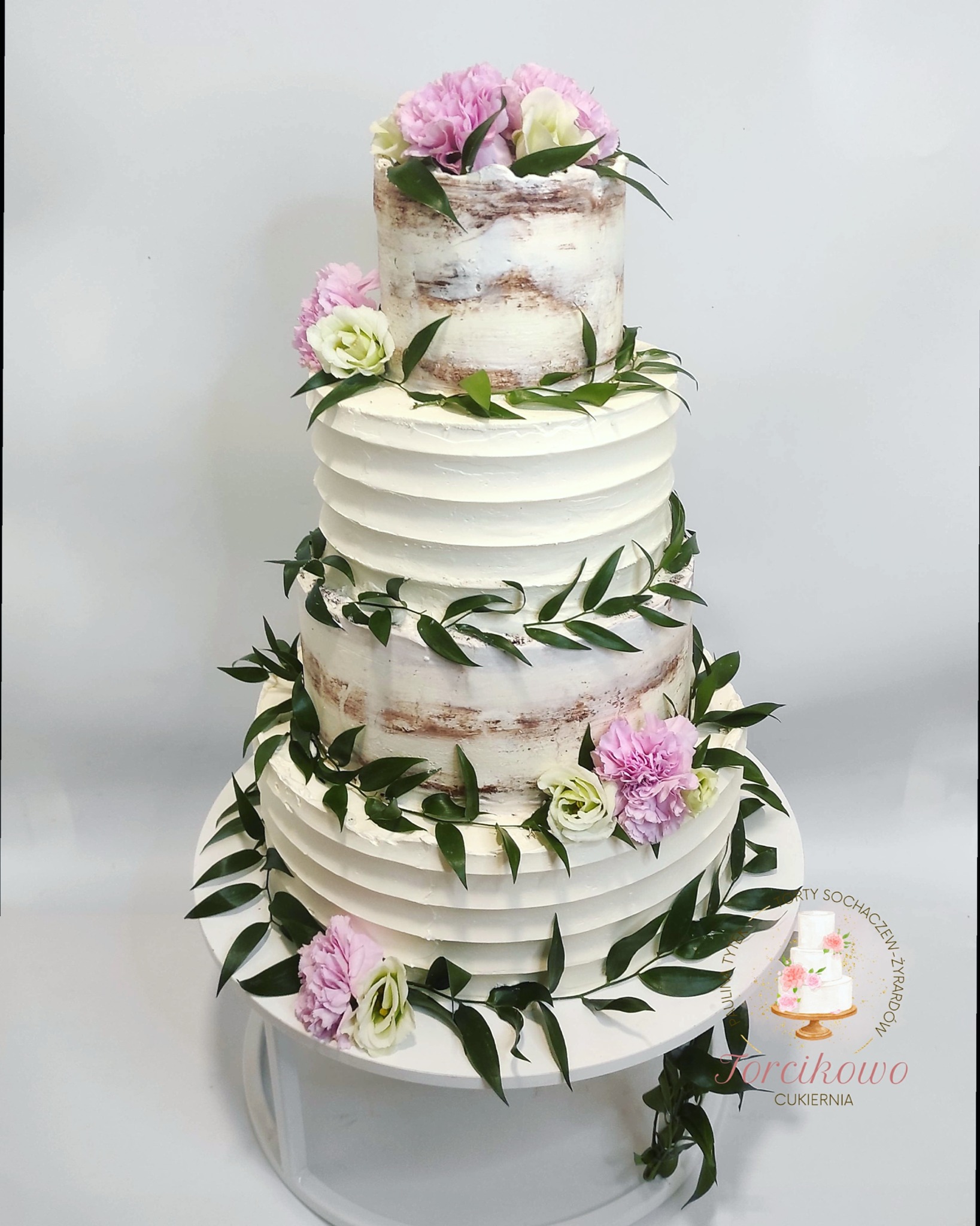 Trzypiętrowy tort weselny w stylu rustykalnym, udekorowany świeżymi kwiatami i zielonymi liśćmi na białym tle, z widocznym logo na podstawie.