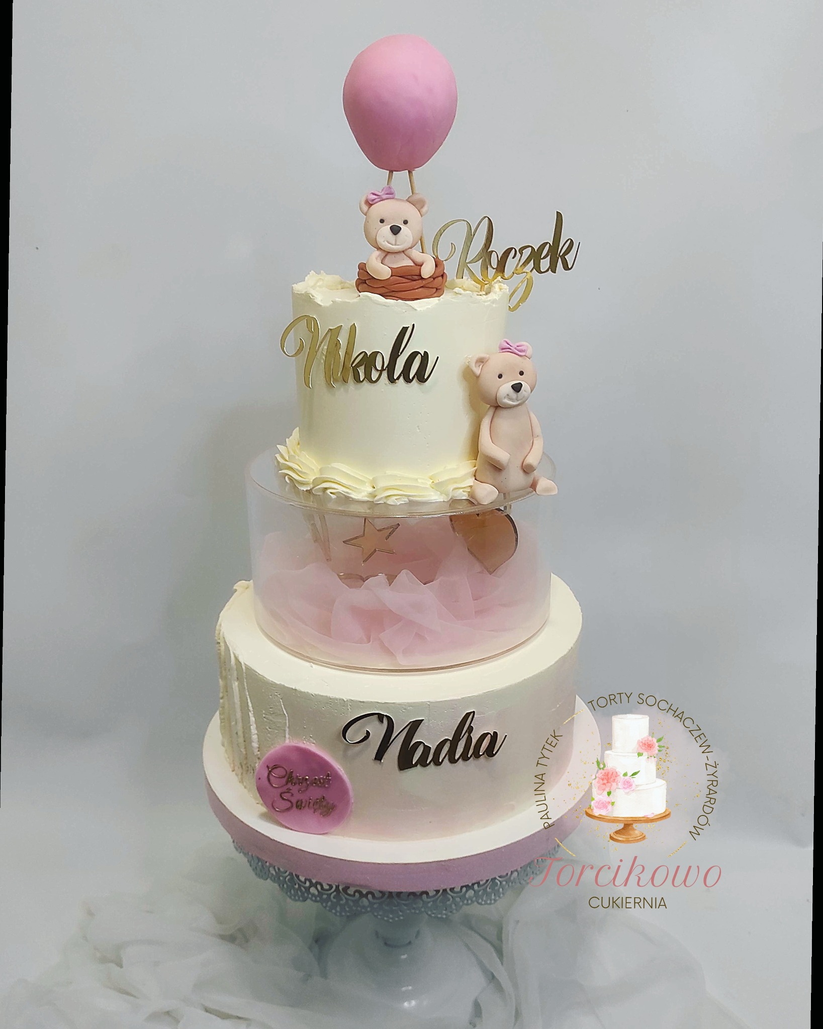 Trzypiętrowy tort urodzinowy w odcieniach bieli i różu, z dekoracjami w postaci figurek misiów, złotych napisów z imionami i cyfrą 'Roczek', oraz różowego balona na szczycie. Środkowe piętro...