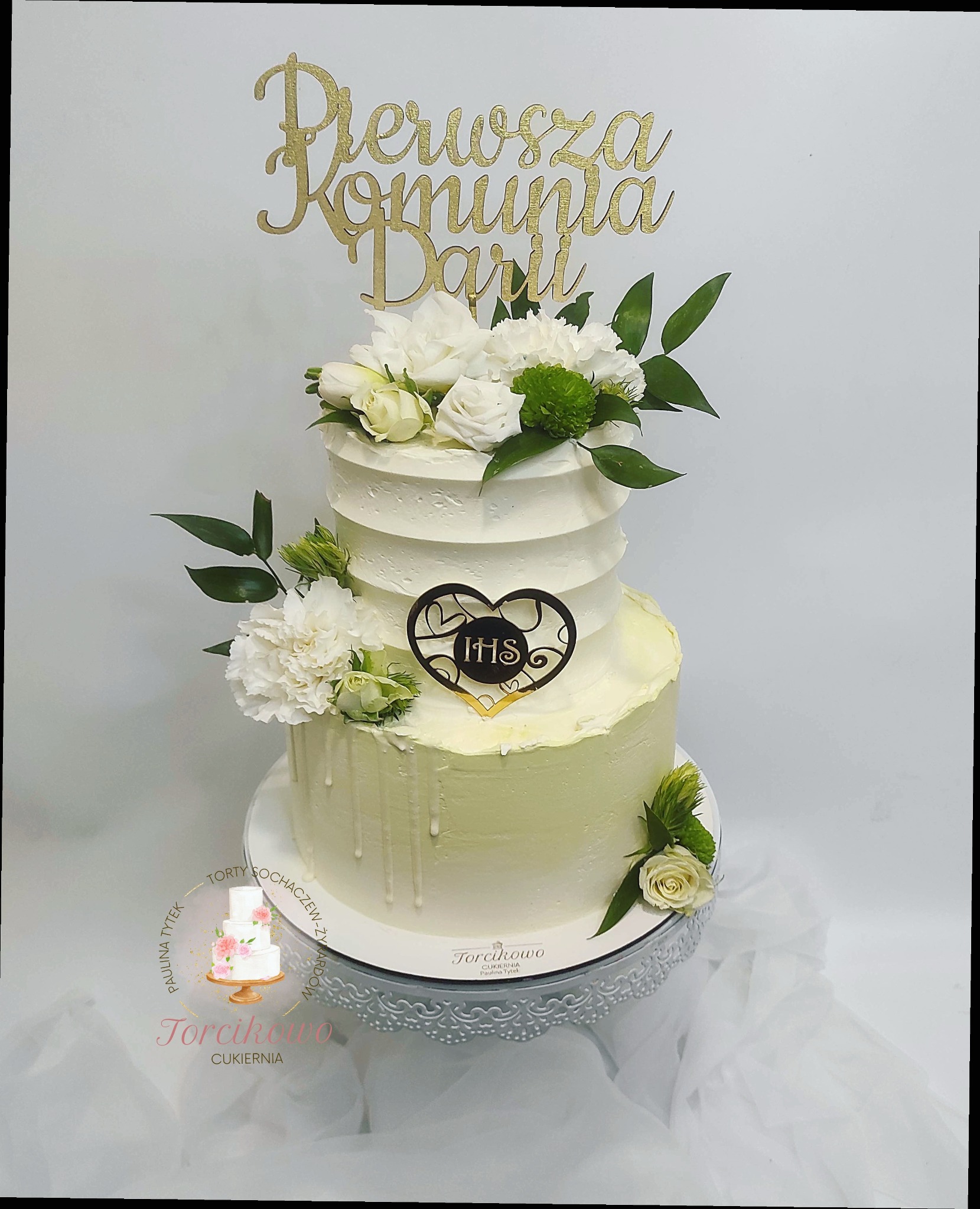 Dwupiętrowy tort z kremem w odcieniach bieli i zieleni, udekorowany białymi kwiatami, zielonymi liśćmi i złotym napisem 'Pierwsza Komunia Darii' oraz sercem z symbolem IHS, na srebrnej paterze.